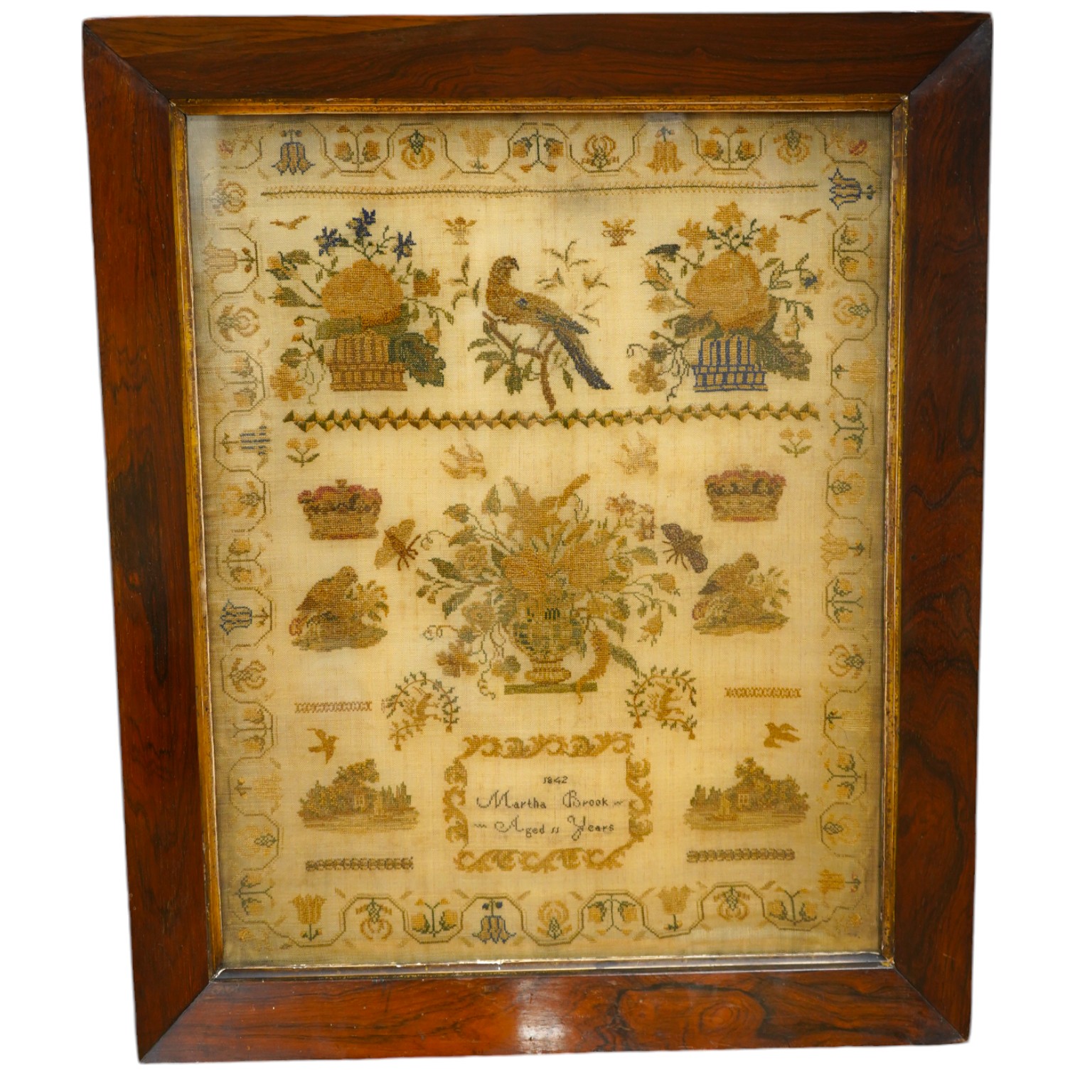 A rosewood framed sampler, dated 1842, by Martha Brook, aged 11years, worked above with large motifs of flower baskets and a bird, a vase of flowers beneath, surrounded by crowns, butterflies and chicks above two small c